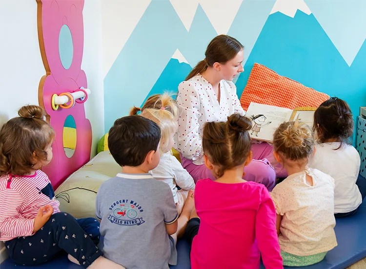 Une assistante maternelle de crèche lit une histoire à un groupe de jeunes enfants assis sur des coussins